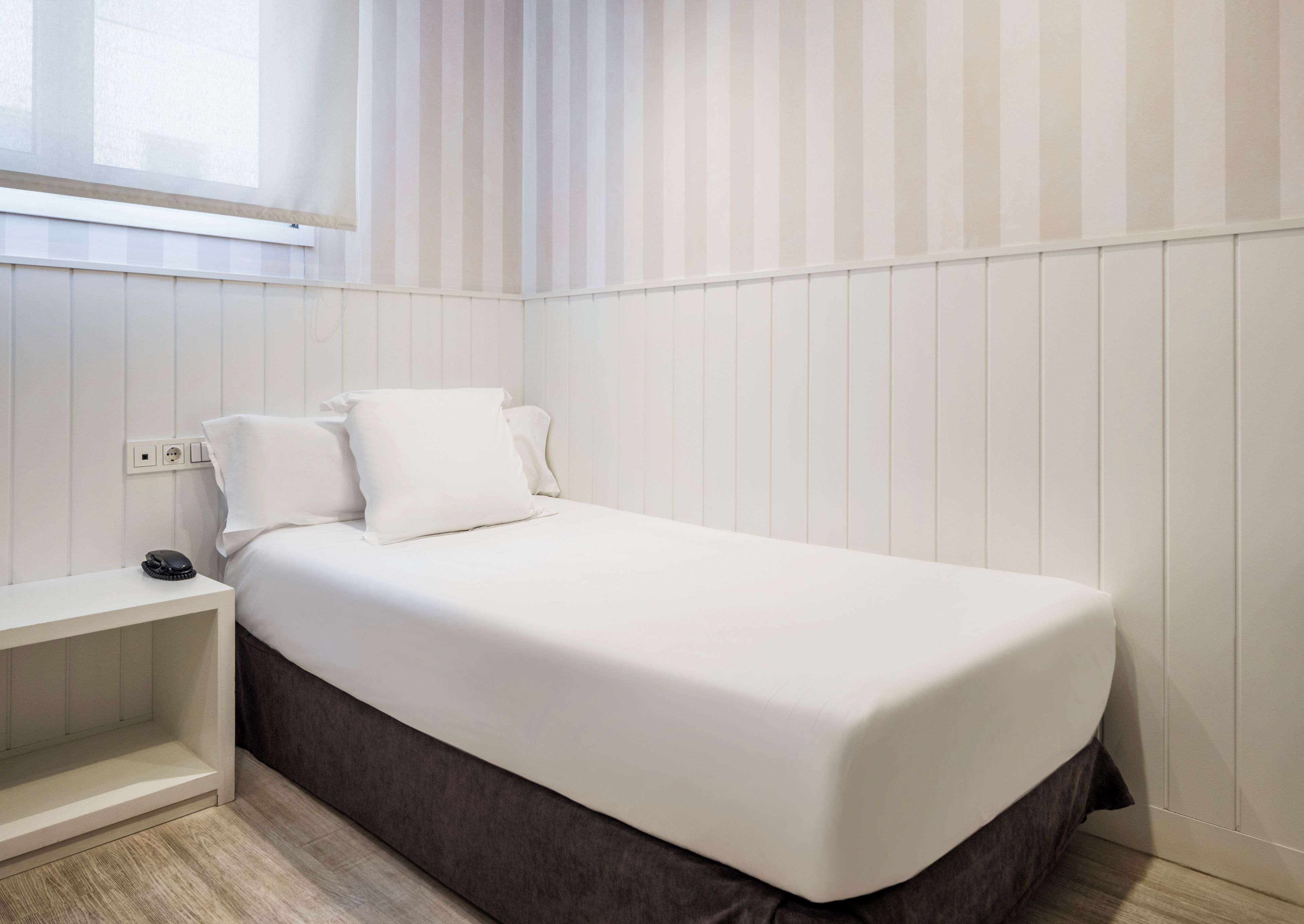 A neatly made bed with white bedding in a room with white paneled walls and a window with vertical blinds.