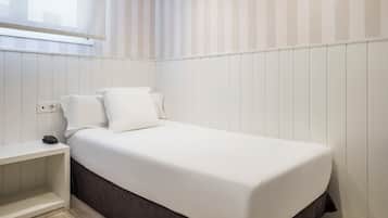 A neatly made bed with white bedding in a room with white paneled walls and a window with vertical blinds.
