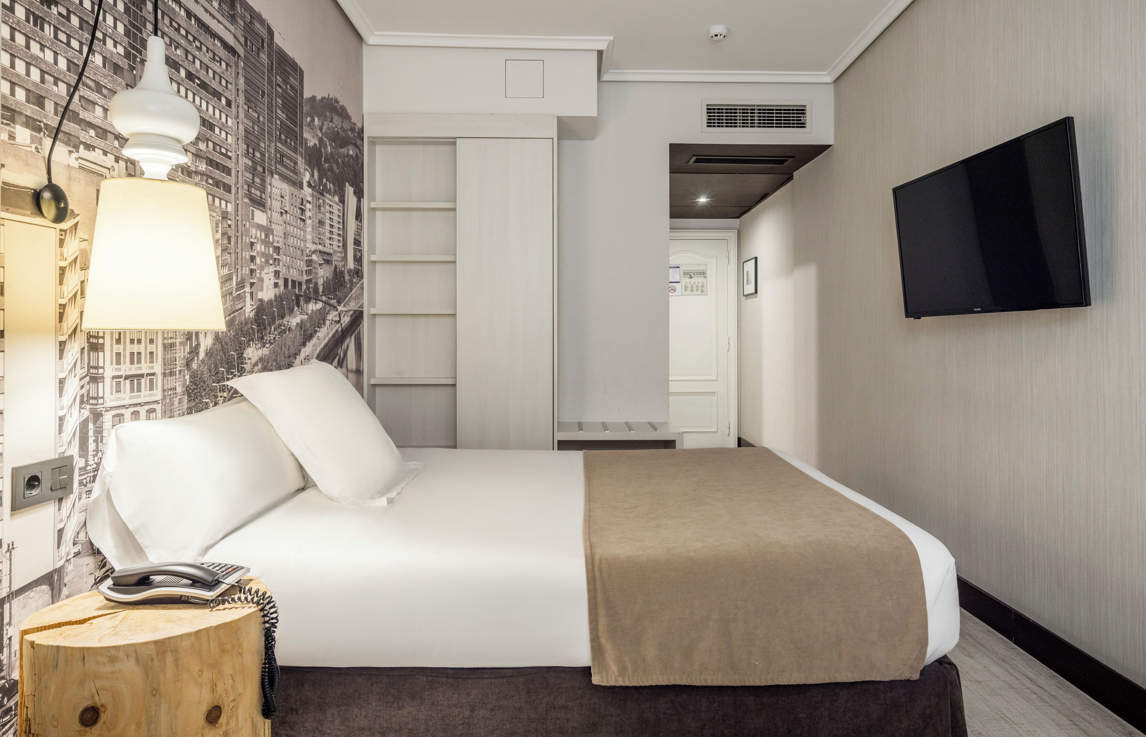 Habitación de hotel con cama, televisor de pantalla plana montado en la pared, mesita de noche de madera con teléfono y lámpara de pared.