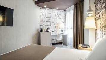 A hotel room with a bed, a desk with a chair, a bookshelf, a lamp, and a telephone on a wooden stool.
