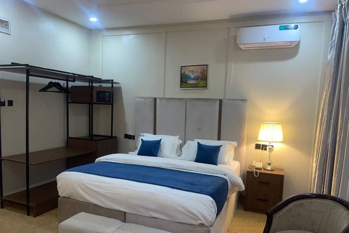 A hotel room with a bunk bed, a single bed, a bedside table with a lamp, a chair, and an air conditioning unit.