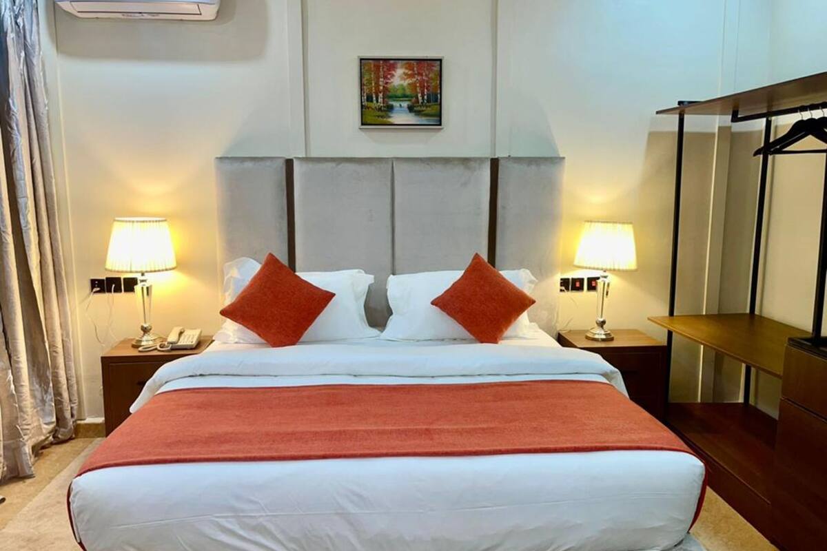 A hotel room with a bed, two bedside lamps, a painting on the wall, and a coat rack.