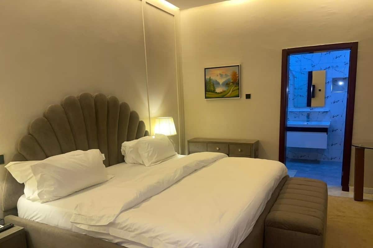 A hotel room with a large bed, a nightstand, a bedside table, a lamp, a painting on the wall, and a bathroom visible through an open door.