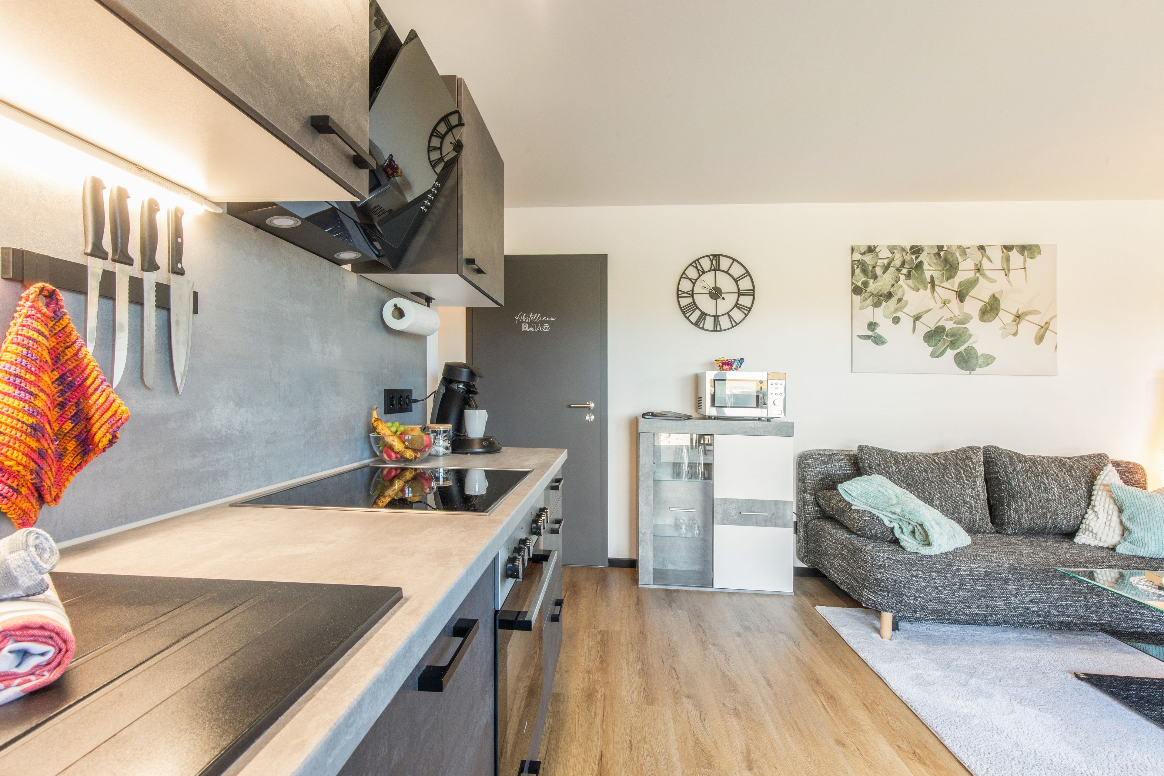 Eine moderne Küche mit Einbauherd, Mikrowelle und Kaffeemaschine. Es gibt einen Essbereich mit einem grauen Sofa und einem kleinen Tisch.