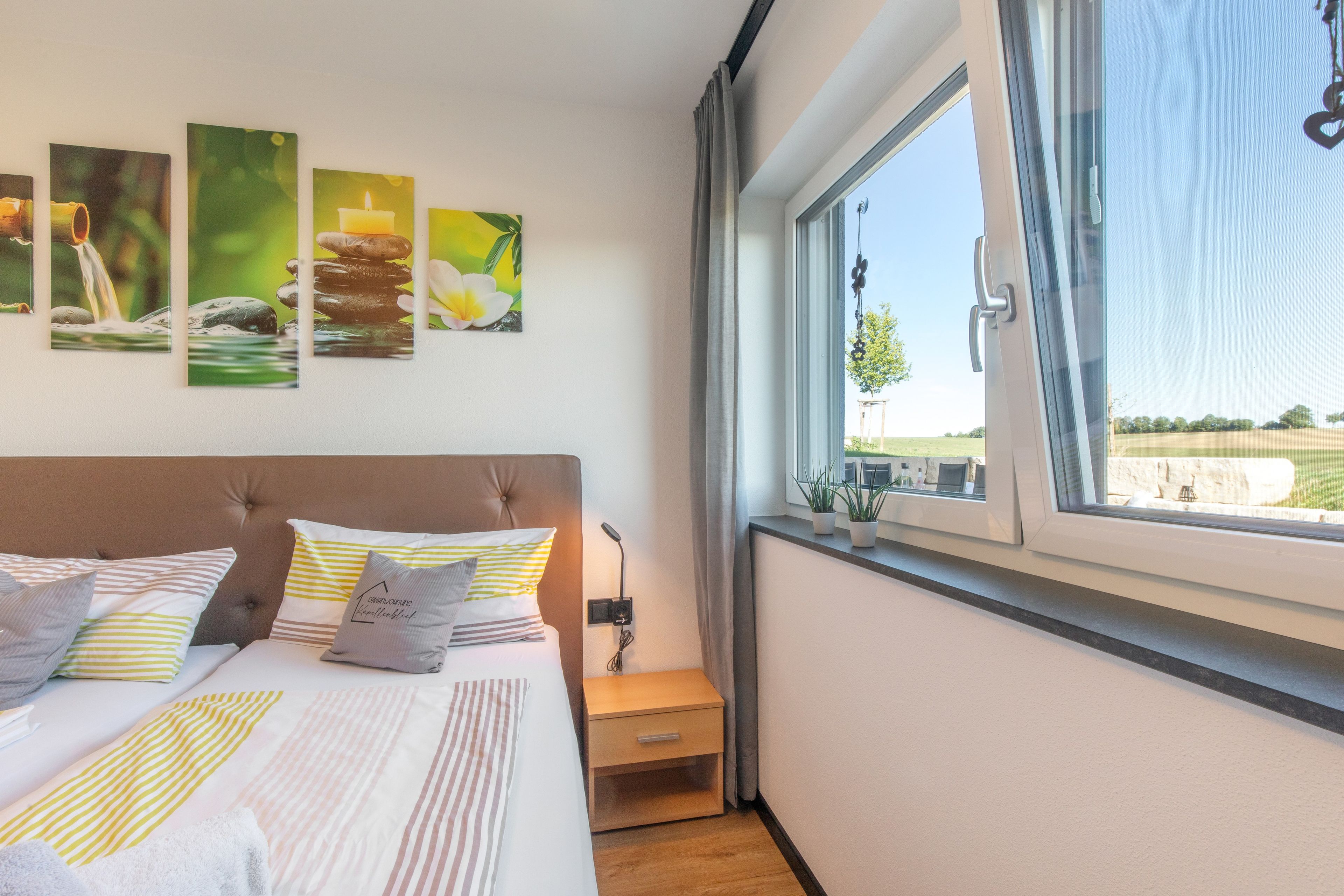 Ein Schlafzimmer mit Bett, Kissen, einem Nachttisch, einem Fenster und Wandbildern.