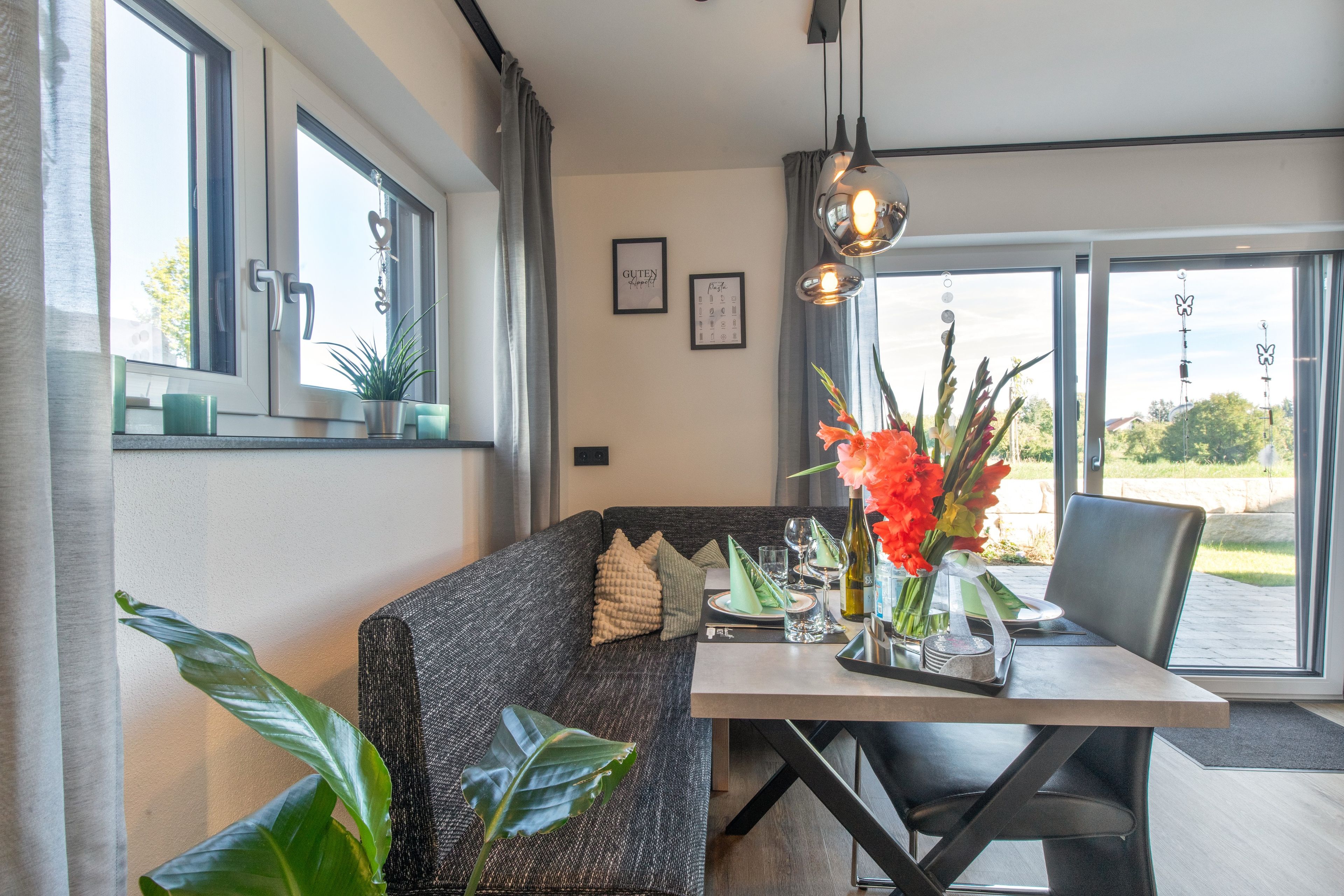Uma sala de jantar moderna com sofá, mesa e cadeiras. Há plantas em vasos e uma janela com vista.
