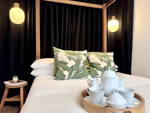 Ein Bett mit Baldachin, zwei Kissen mit Blattmuster, ein Tablett mit weißen Teetassen und einer Teekanne, sowie ein kleiner Holzschemel mit einer Pflanze.