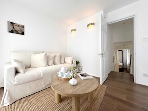 Ein minimalistisch eingerichtetes Wohnzimmer mit einer Couch, einem runden Holztisch und einer Tür zu einem weiteren Raum.