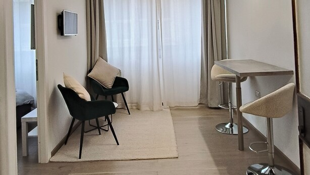 Ein modernes Hotelzimmer mit Bett, Schreibtisch, zwei Stühlen, einem kleinen Tisch, Fernseher und einem Fenster mit Vorhängen.