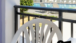 Un balcón con una silla blanca, una piscina con sillas de descanso y sombrillas, y un edificio al fondo.