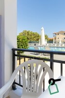 Un balcone con una sedia bianca, una piscina con lettini e ombrelloni, e un edificio sullo sfondo.