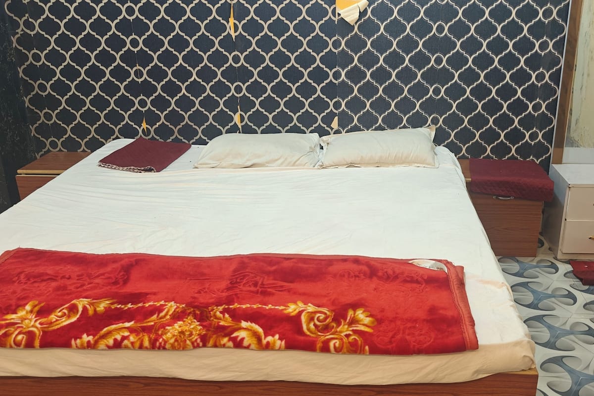 A neatly made bed with a red blanket, a bedside table with a drawer, and a patterned wall behind the bed.