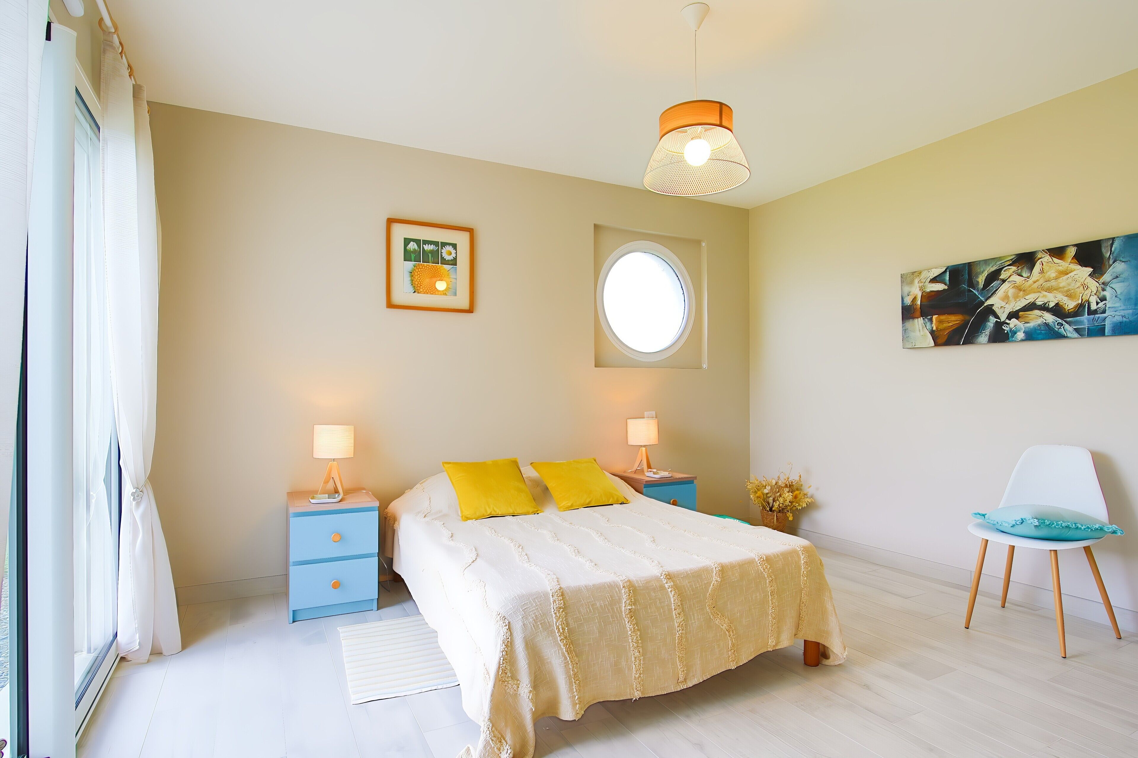 Una camera da letto con un letto, due comodini con lampade, una finestra, uno specchio rotondo, una sedia e opere d'arte alle pareti.