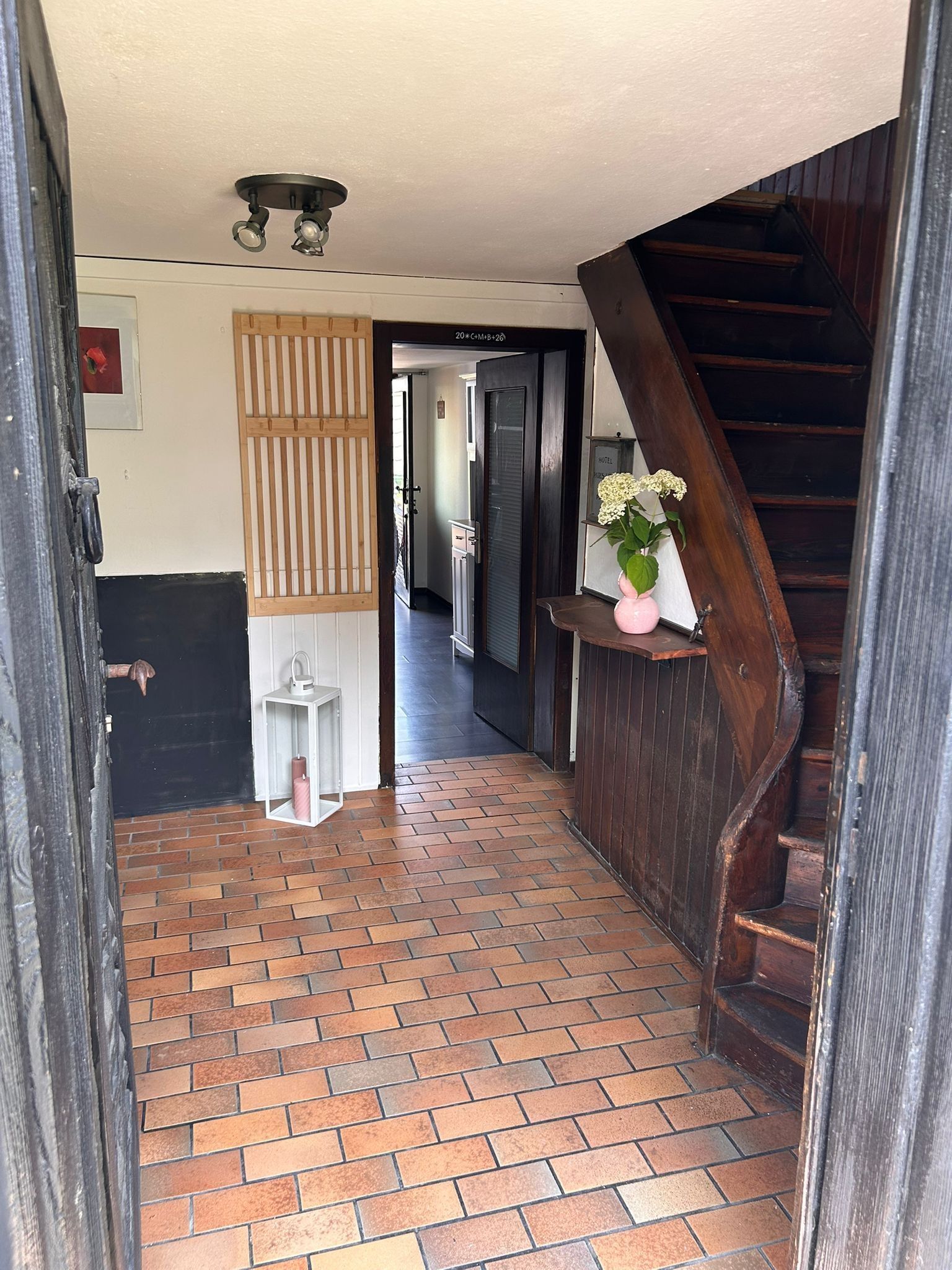 Un pasillo con suelo de ladrillo, una puerta de madera, un jarrón blanco con flores y una escalera.