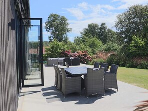 Um pátio externo com mesa e cadeiras de jantar, porta de vidro que dá para um jardim e vegetação exuberante ao fundo.