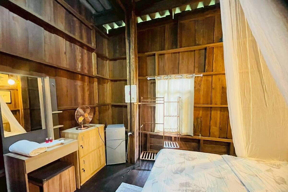 A rustic wooden room with a bed, a small desk with a mirror, a fan, and a shelf with a ladder.
