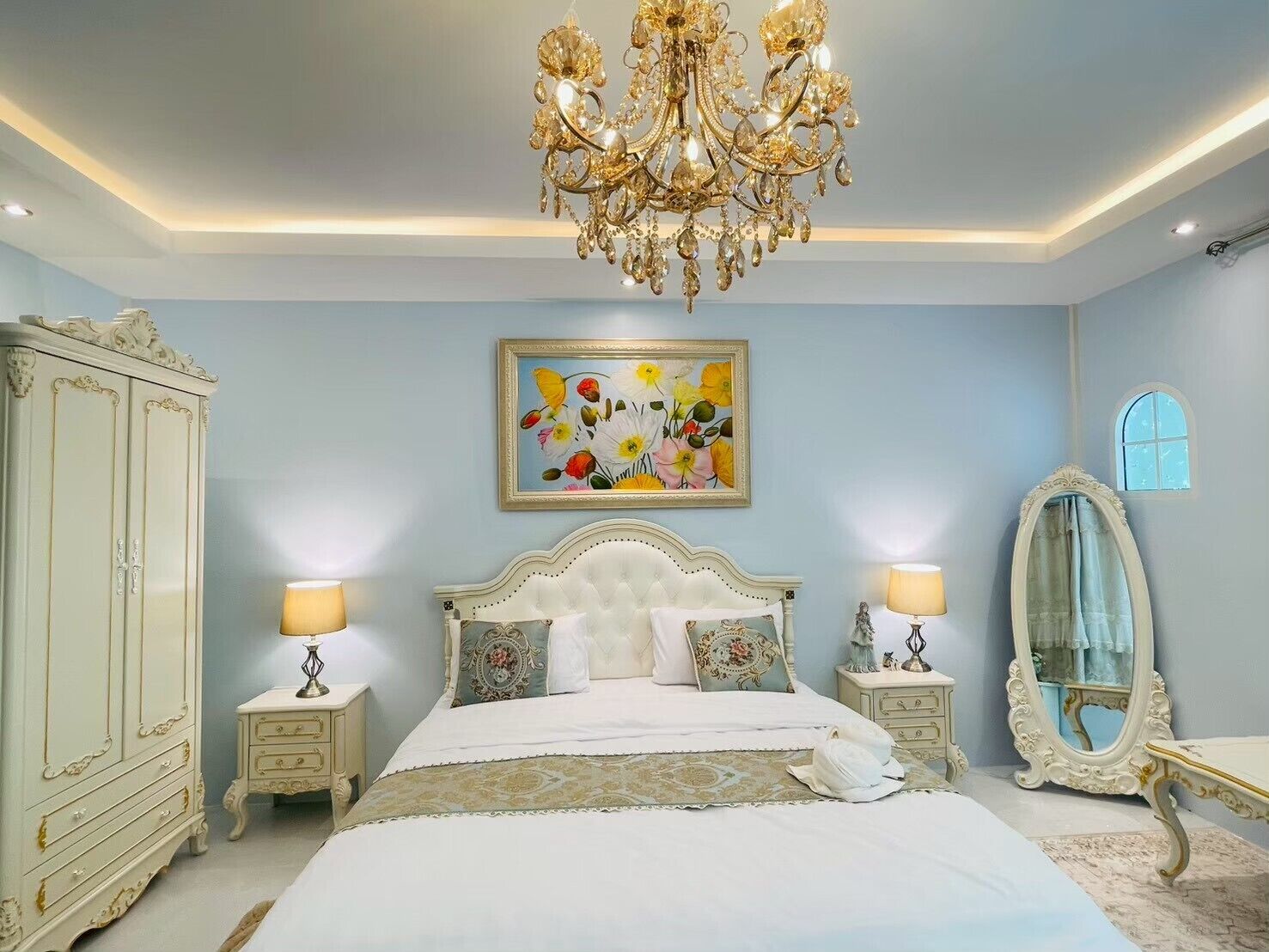 A bedroom with a large bed, a chandelier, a wardrobe, a mirror, and a floral painting on the wall.