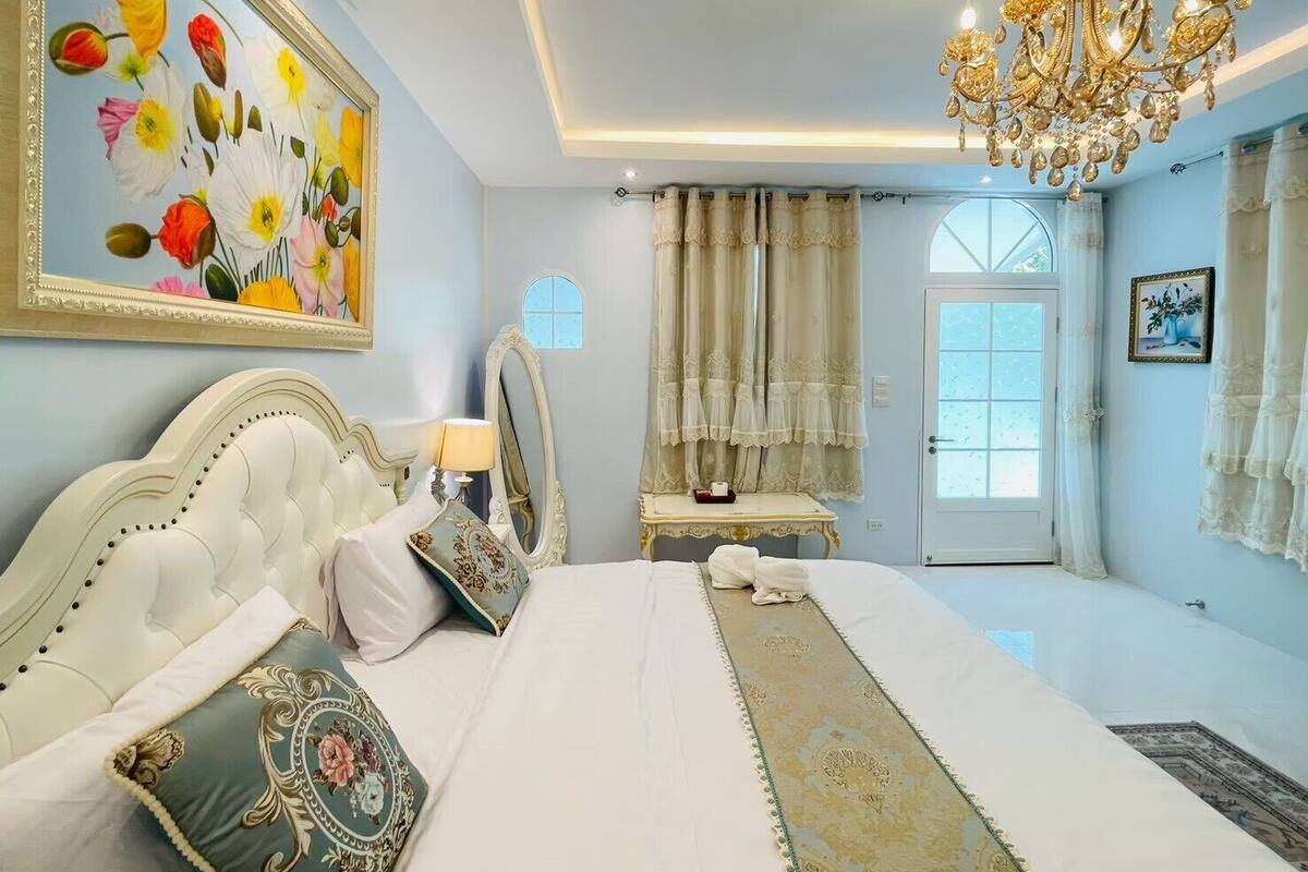 A bedroom with a large bed, a chandelier, a painting, and a window.
