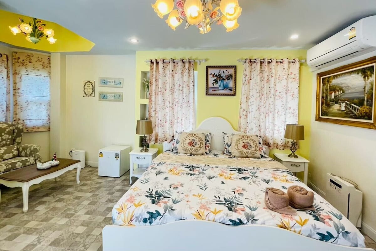 A bedroom with a bed, two bedside tables, a chair, a small table, a ceiling light fixture, and an air conditioning unit.