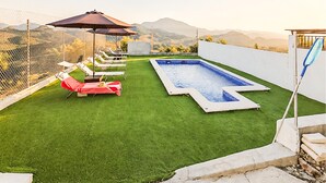 A backyard with artificial grass, a swimming pool, lounge chairs, and a large umbrella.