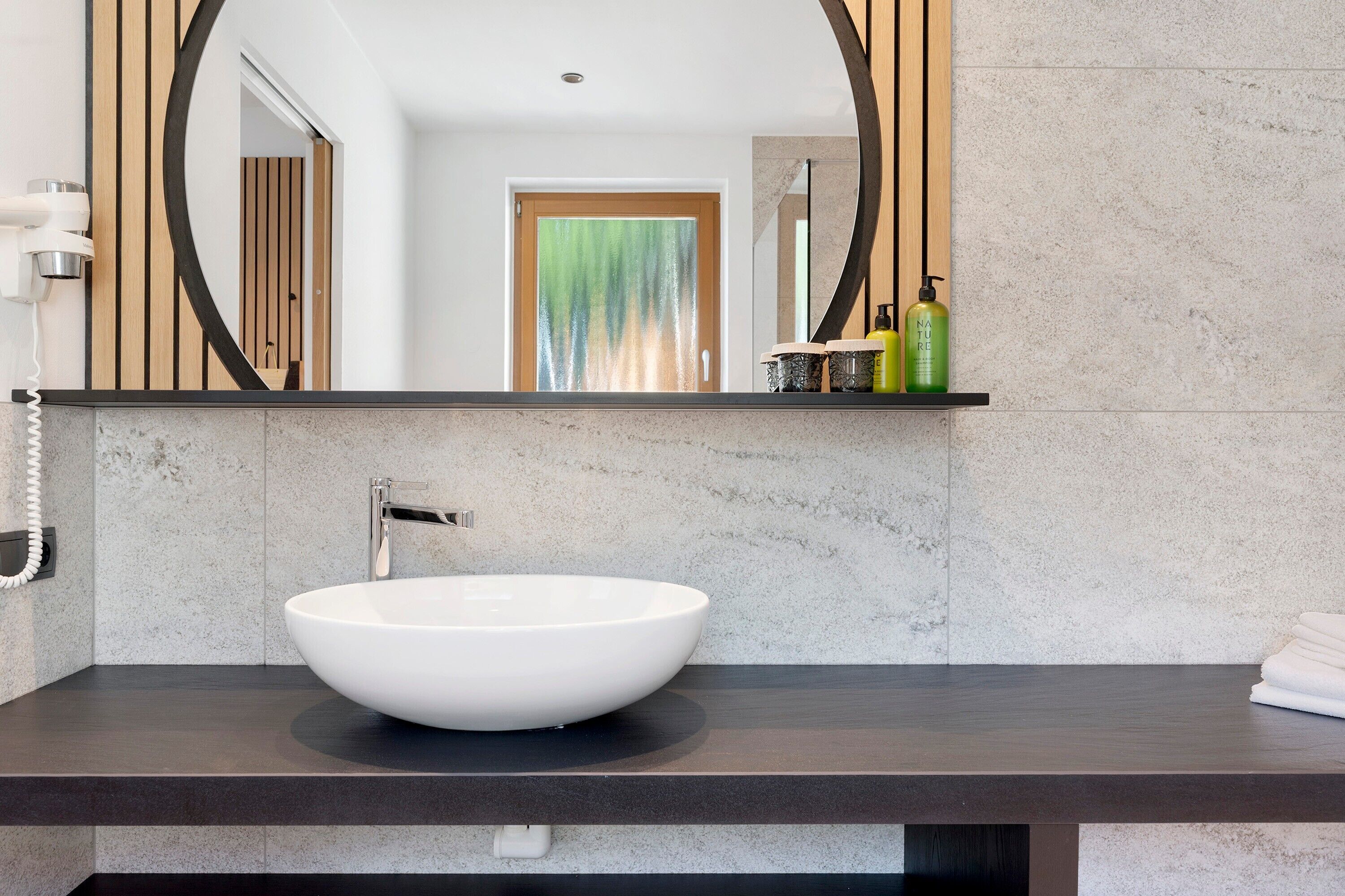 A modern bathroom with a large white vessel sink, a dark countertop, a mirror with a wooden frame, and a towel rack.