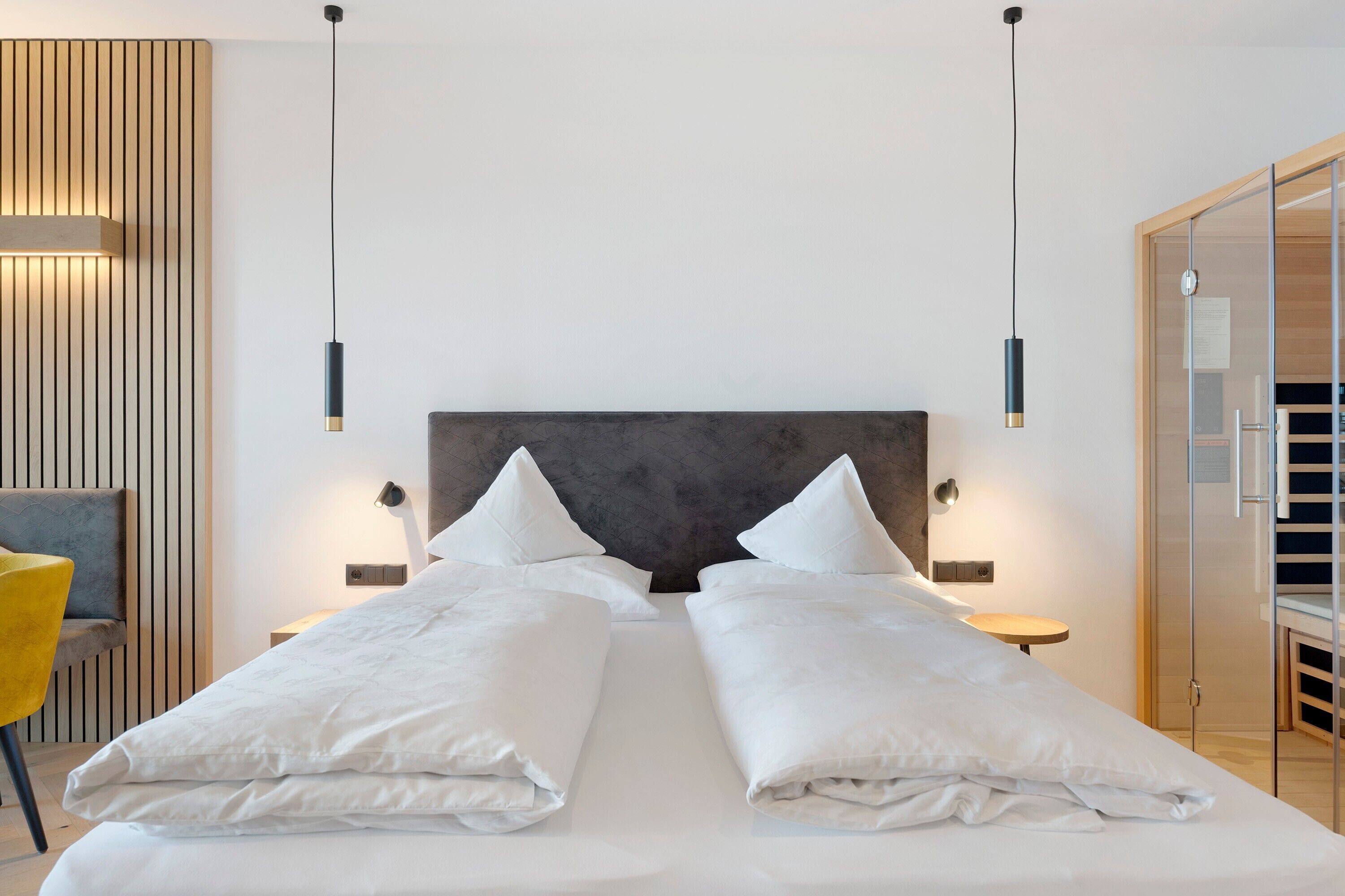 Quarto de hotel com uma cama grande, mesas de cabeceira, uma cadeira e uma porta de vidro para um banheiro.