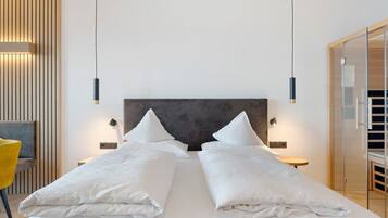 Quarto de hotel com uma cama grande, mesas de cabeceira, uma cadeira e uma porta de vidro para um banheiro.