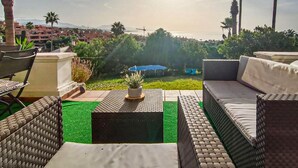 Un área de estar al aire libre con sofá, sillas y una mesita, con vista a un jardín y a edificios y montañas.