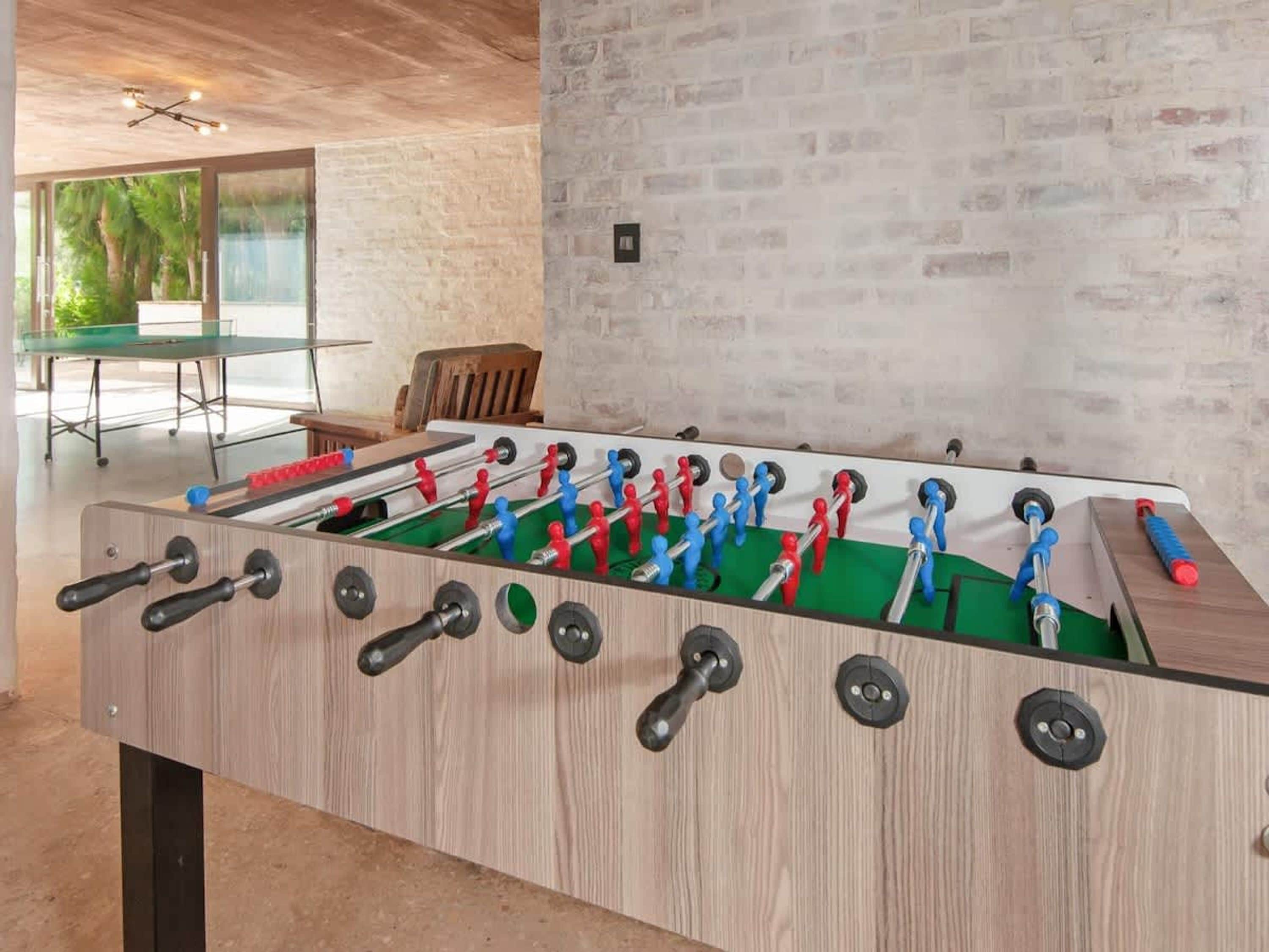 A wooden foosball table in a room with a brick wall, a glass table, and a wooden chair.