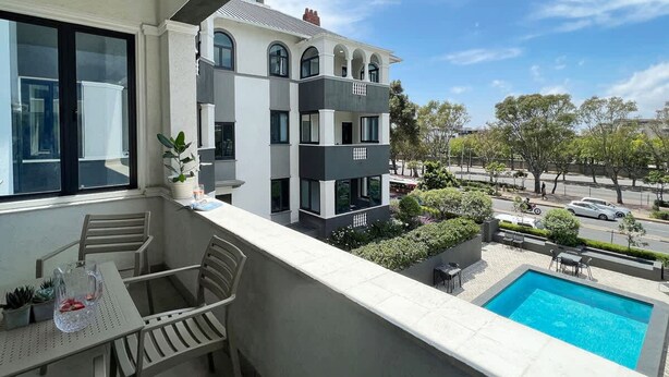 Uma varanda com piscina, mesa e cadeiras, e vista de um prédio com vários andares.