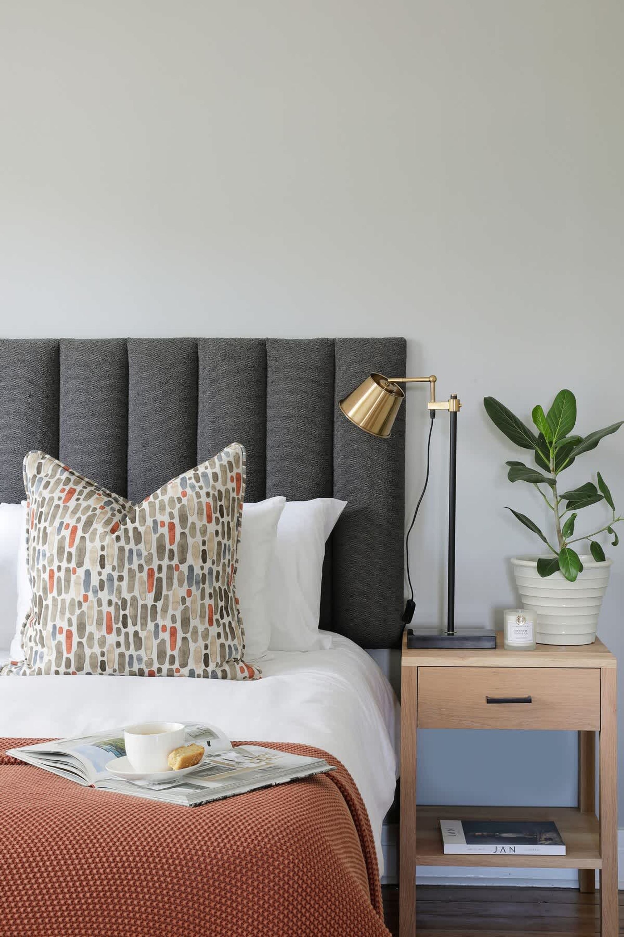 Una cama bien hecha con un cojín estampado, una lámpara de noche, una planta en maceta y una mesita de noche con un libro.