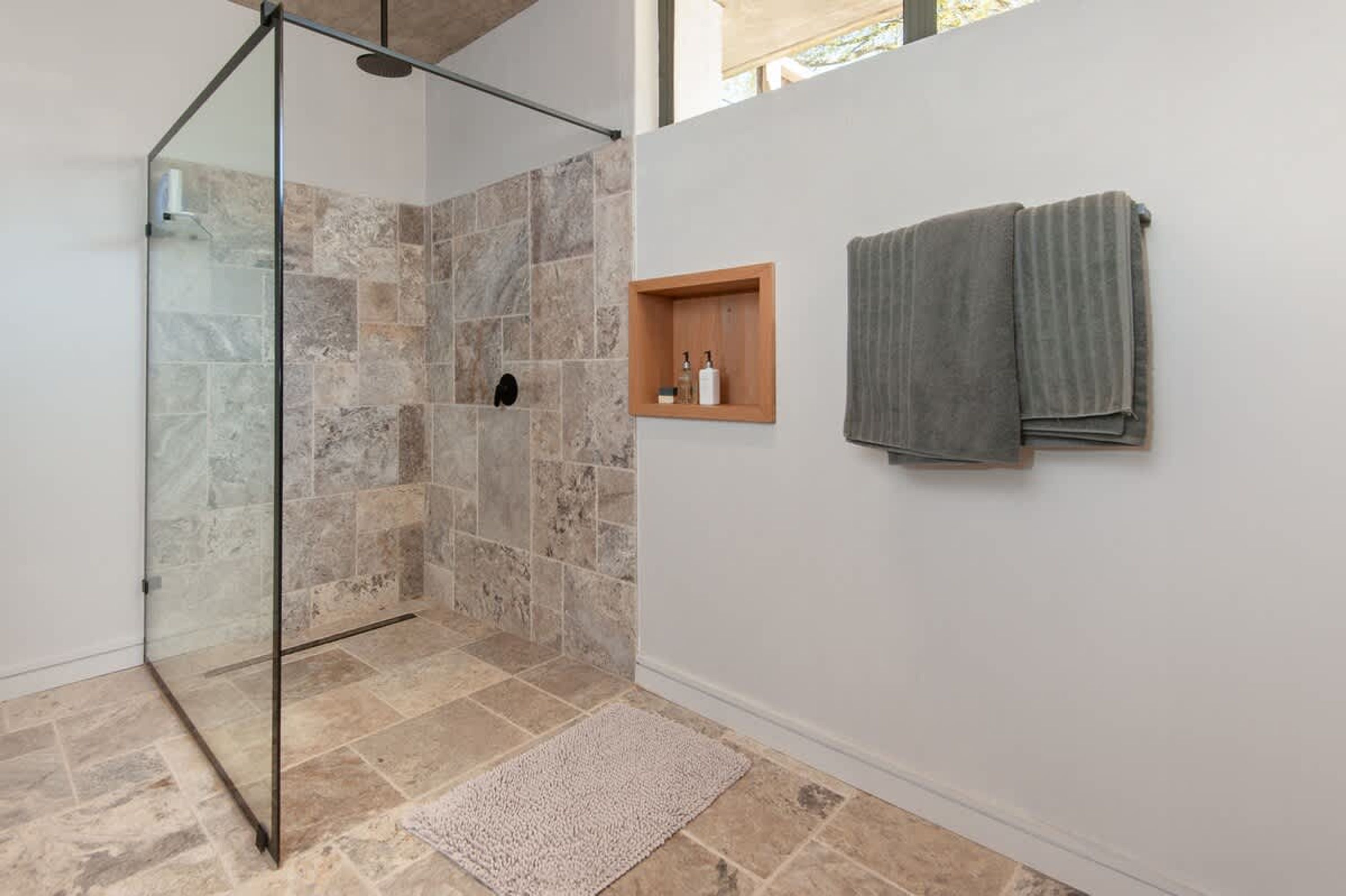Ducha con puertas de vidrio, estante de madera con artículos de aseo y un perchero en una pared blanca.