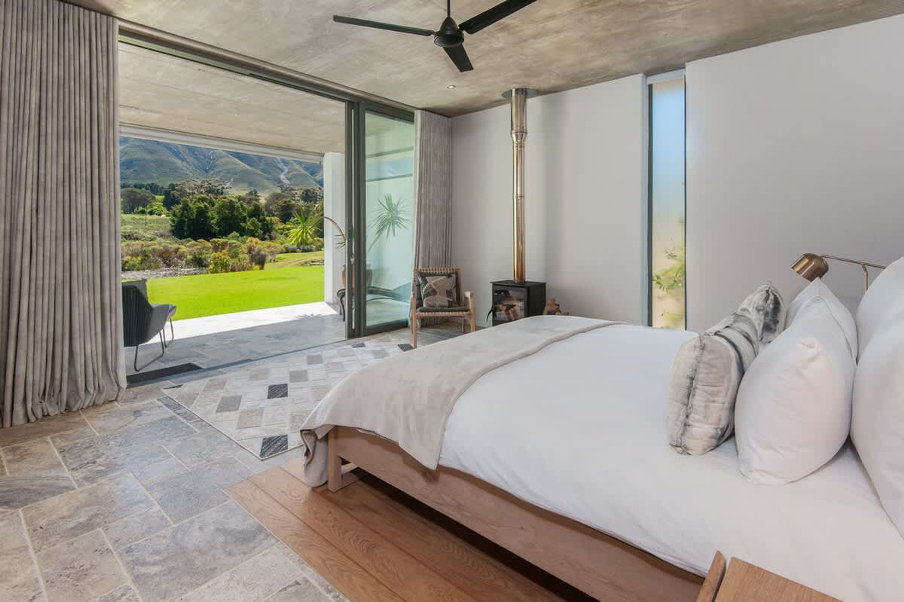 Quarto com uma cama grande, ventilador de teto, lareira e vista para uma paisagem verde através de uma porta de correr de vidro.