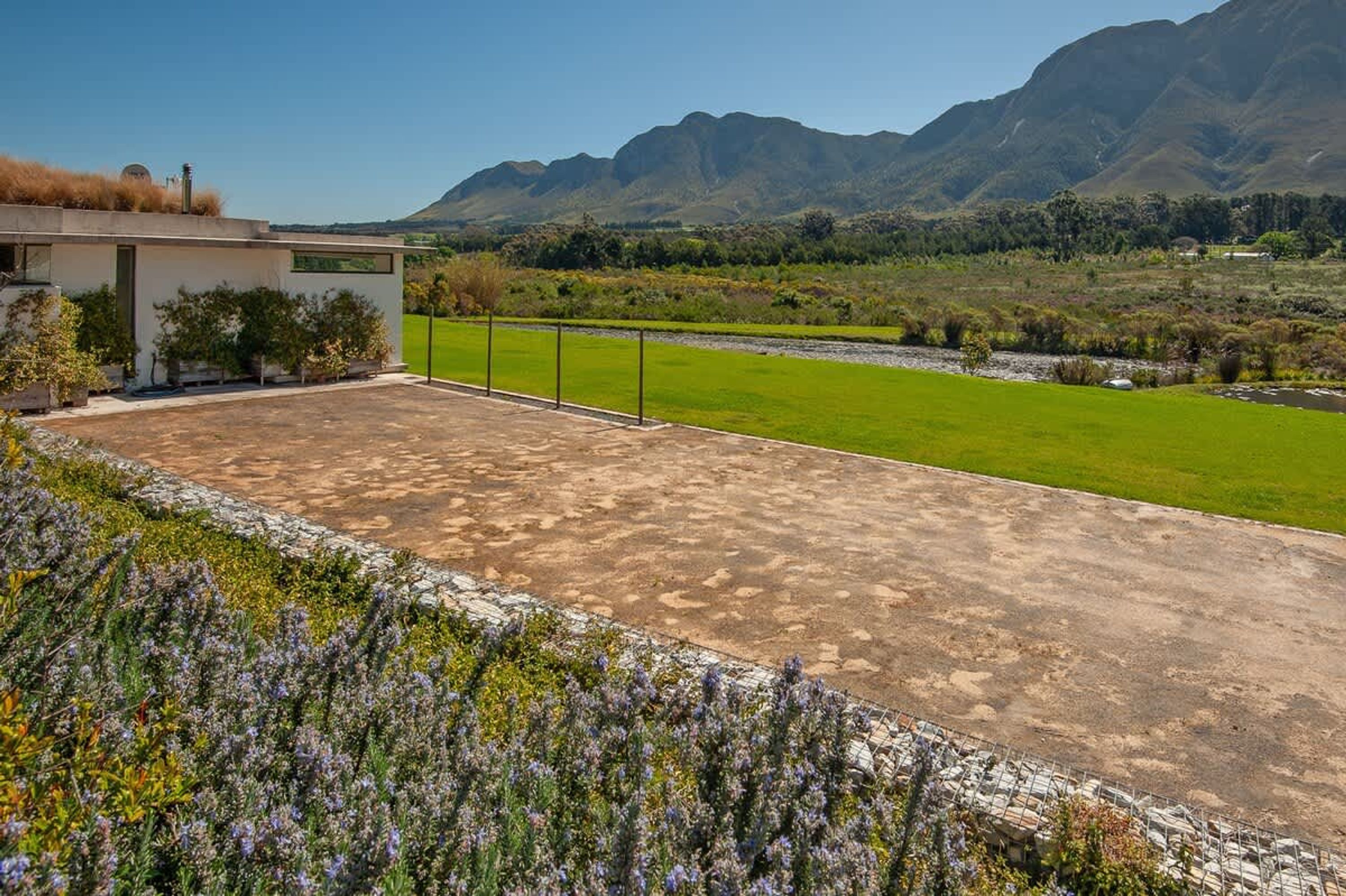 A modern building with a flat roof, a paved patio, and a view of a green field and mountains.
