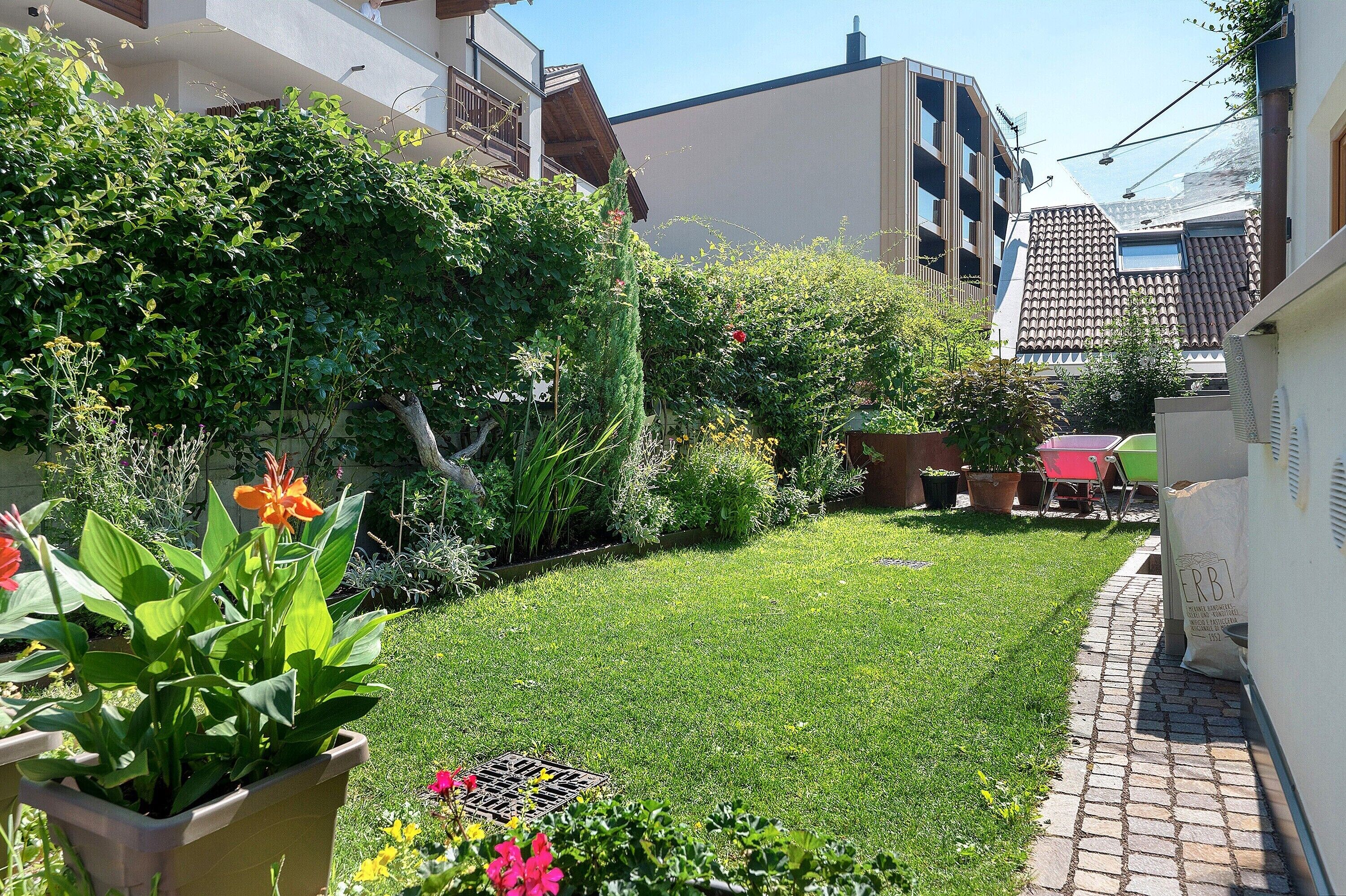 Ein schmaler Stadtgarten mit gepflastertem Weg, Blumentöpfen und vielfältigem Grün.