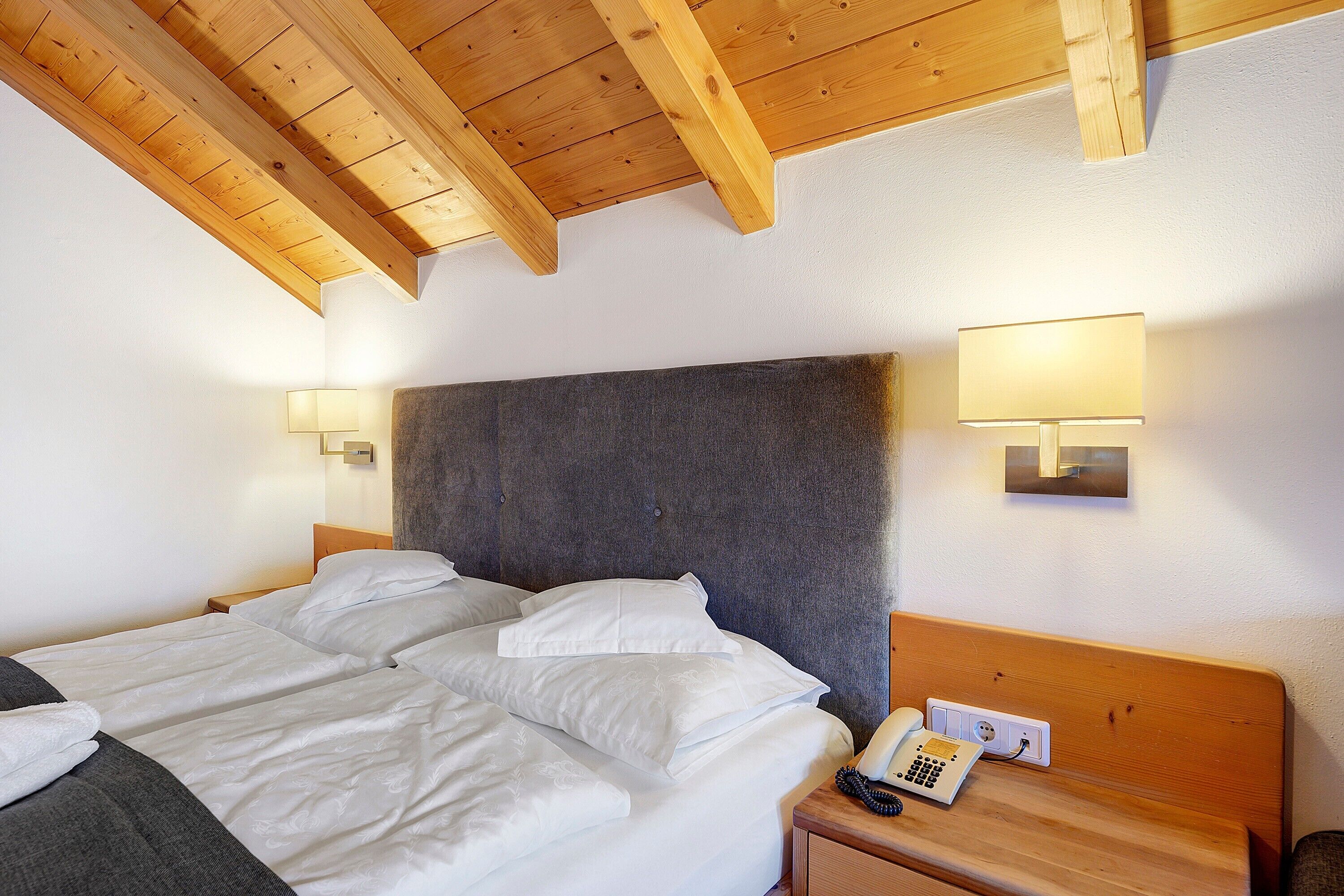 Habitación de hotel con cama, lámparas de noche, teléfono y cabecero de madera.