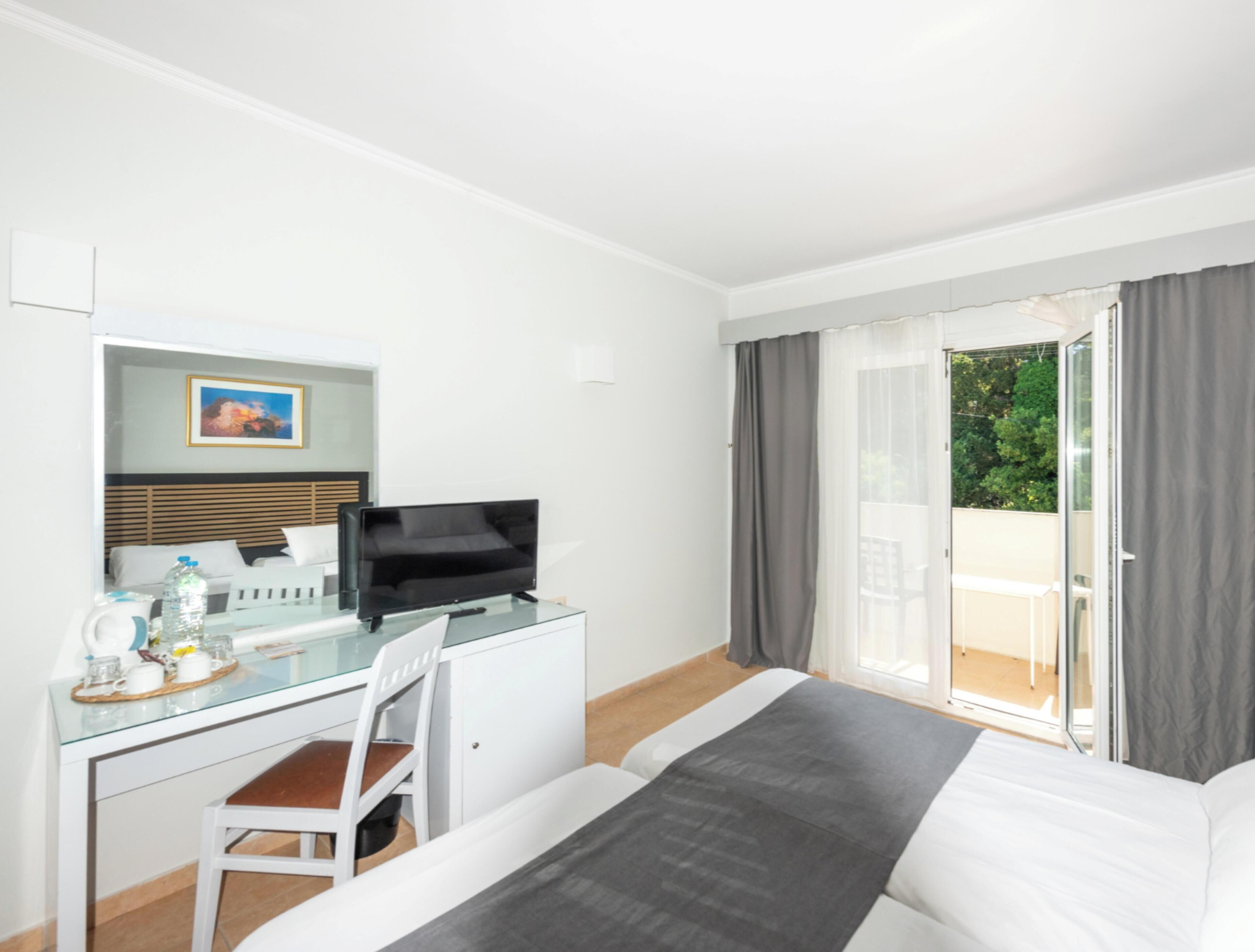 Habitación de hotel con cama, escritorio con ordenador, silla, ventana con cortinas y vistas a zonas verdes.