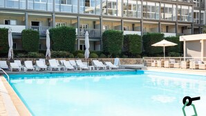 Una piscina con sillones de descanso y sombrillas, rodeada por un edificio de varios pisos con balcones.