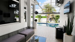 Un espacio exterior moderno con mesa de vidrio, sofá de mimbre, ventilador de techo, televisor y vistas a una piscina y follaje.