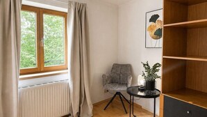 A room with a window, a radiator, a chair, a small table with a plant, and a wooden shelf.