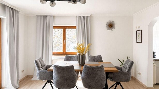 A dining area with a wooden table, grey upholstered chairs, a vase with yellow flowers, and a sunburst wall decoration.