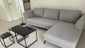 Ein graues Sofa, ein schwarzer Couchtisch und ein kleinerer Beistelltisch in einem Raum mit weißen Wänden und Fliesenboden.