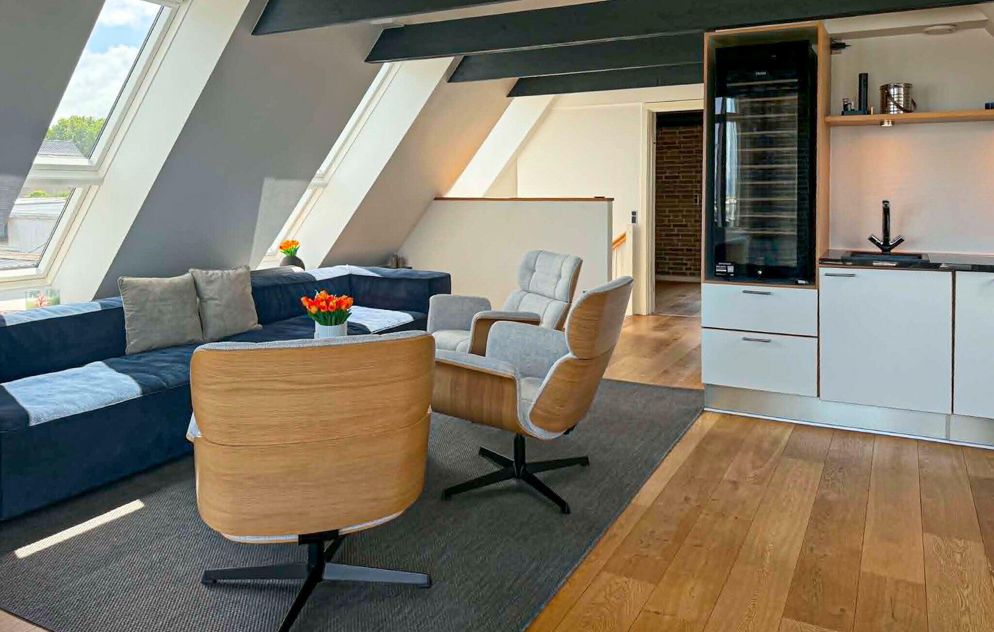 A modern loft apartment with a sloped ceiling, a dark blue sofa, a wooden accent chair, a small table with flowers, and a kitchen area with white cabinets and a black appliance.