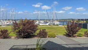 Un puerto deportivo con varios veleros amarrados, una zona verde con arbustos y un cielo despejado.
