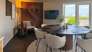 Un comedor moderno con una mesa redonda, seis sillas, un televisor y vistas a barcos a través de un amplio ventanal.