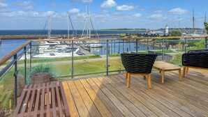 Terraza de madera con mobiliario exterior, con vista a un puerto deportivo con barcos y un faro.