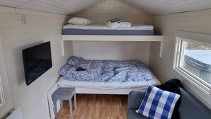 Une petite cabane d’une seule pièce, avec un lit superposé, un canapé gris agrémenté d’un coussin à carreaux, une petite table et un téléviseur fixé au mur.