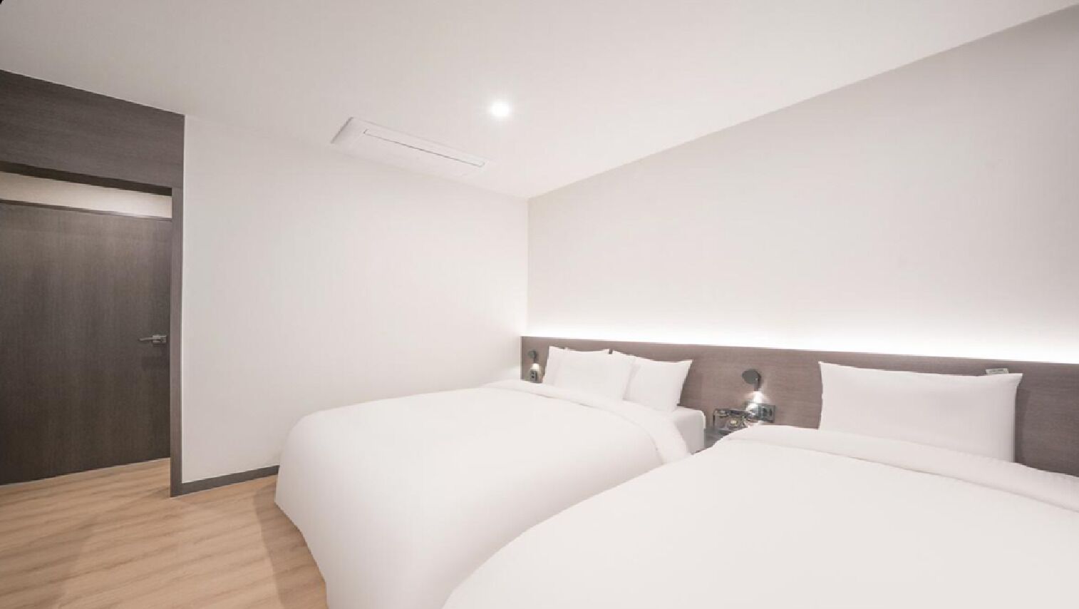 A hotel room with two beds, a wooden floor, a dark door, and a wall-mounted light fixture.