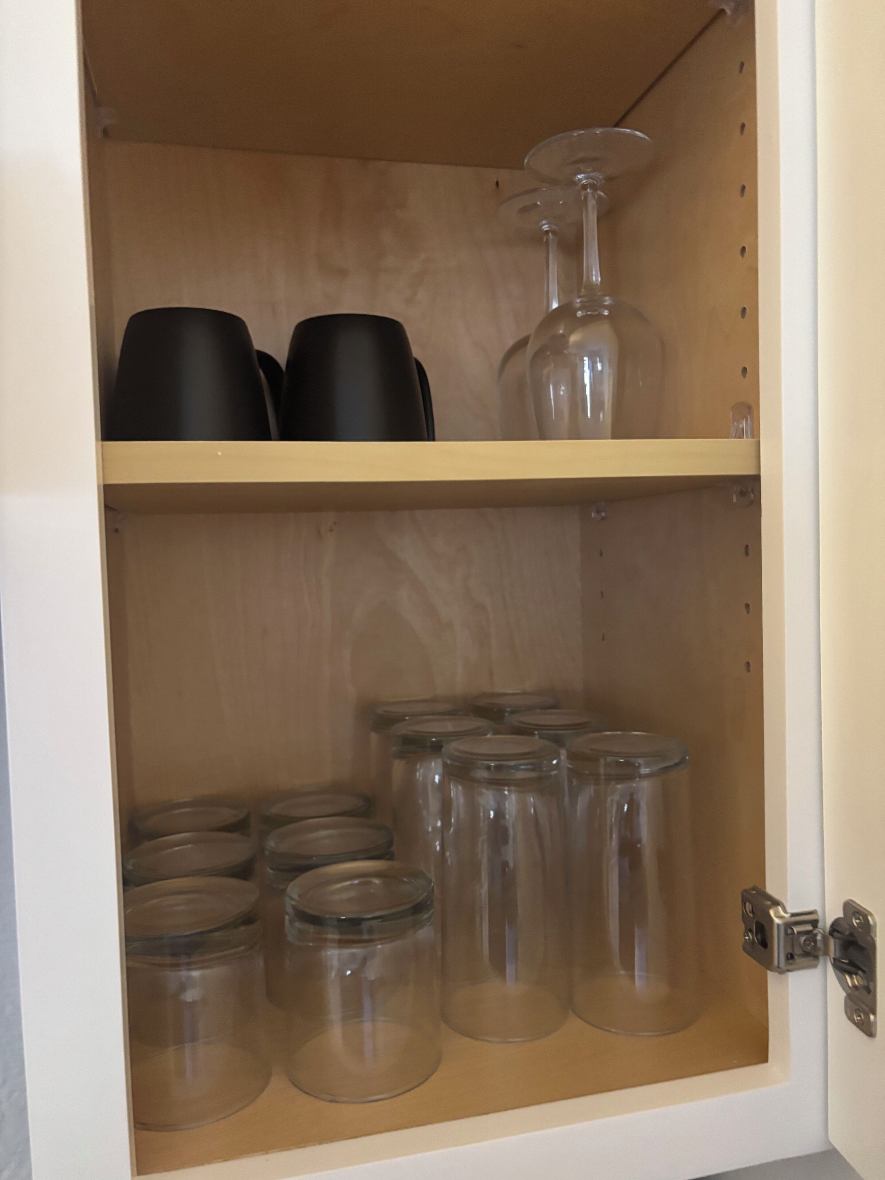 A wooden cabinet with two shelves, one containing black cups and a wine glass, and the other with clear glass jars.