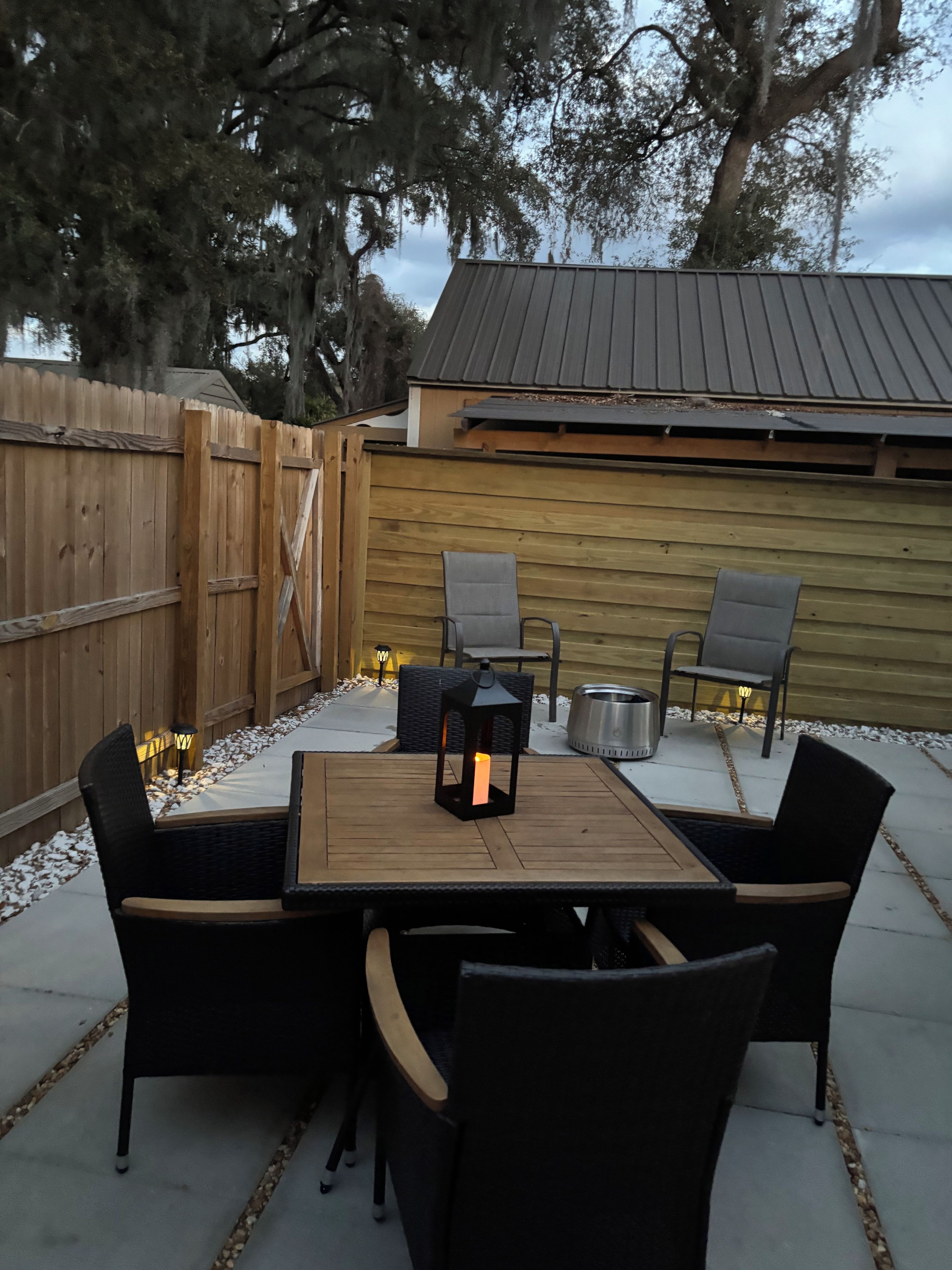 An outdoor patio with a wooden table and four chairs, a lantern, and a metal grill.